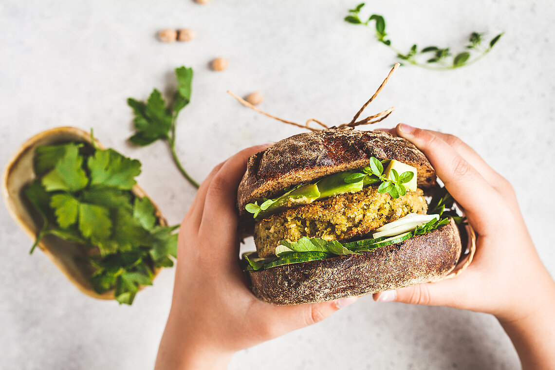ernaehrung-nachhaltig-burger.jpg Kinderhände halten ein veganes Sandwich mit Kichererbsen, Avocado, Gurke und Salat